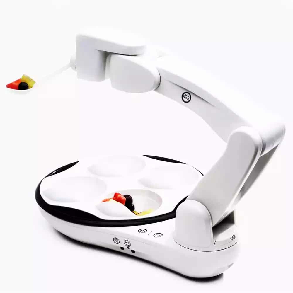 A robotic arm feeder with a spoon attached sits above a divided food tray.
