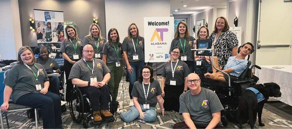 The APTAT team, some using wheelchairs, pose together indoors at an event with a "Welcome! Alabama" sign and various booths in the background.