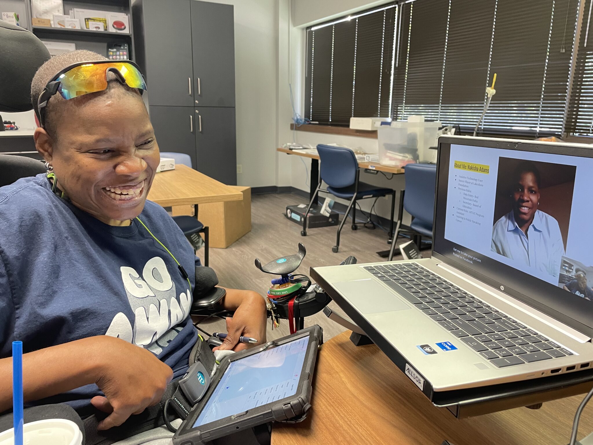 Nakisha smiles while in presentation in an office. She holds a tablet, and her profile is visible on the laptop screen.