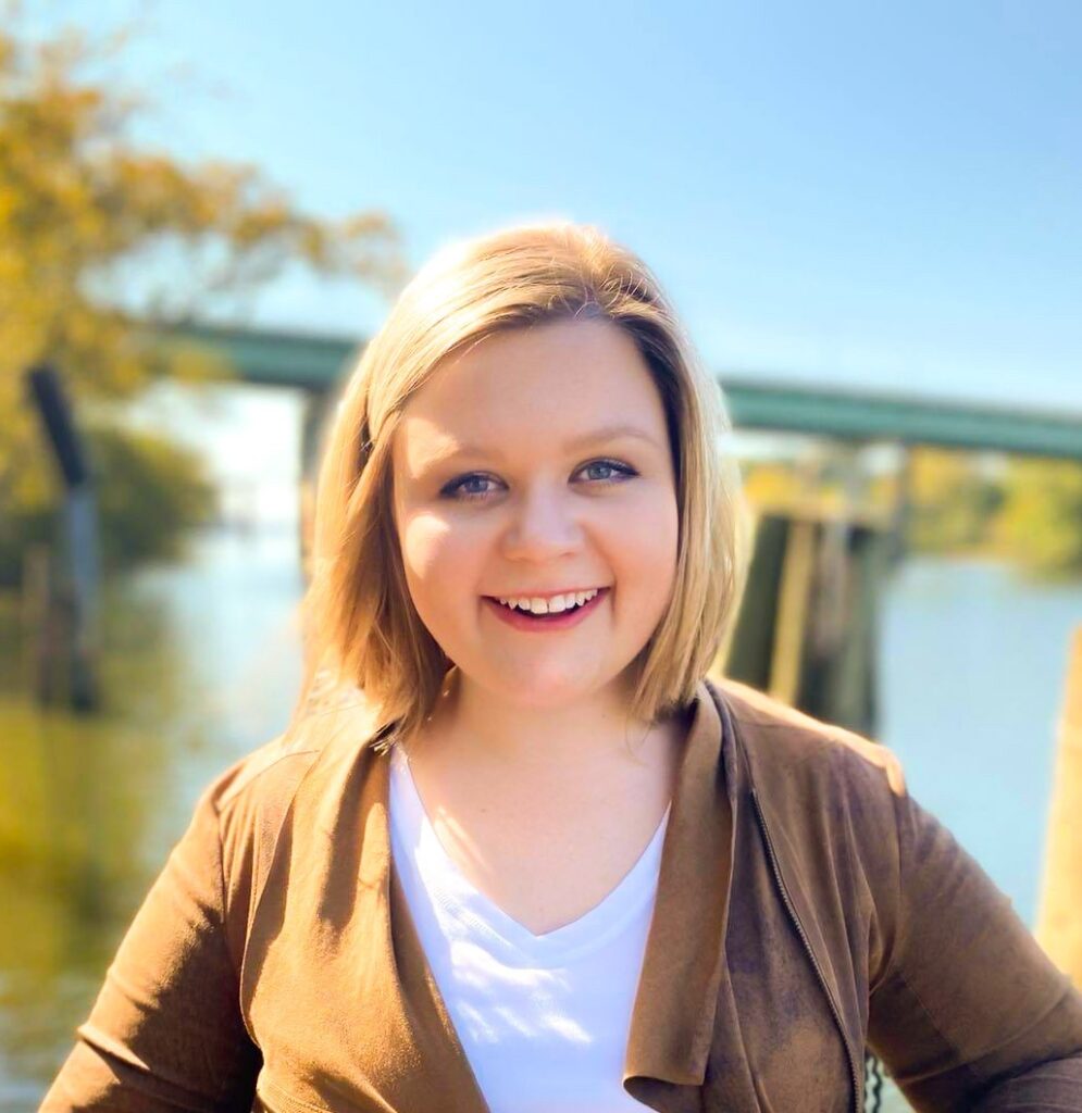 Ashley McLeroy has short blonde hair, smiling outdoors by a river with a bridge and trees in the background on a sunny day.