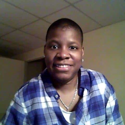 Nakisha Adams wears a blue and white plaid shirt and necklace, smiles at the camera in a room with plain walls and a tiled ceiling.
