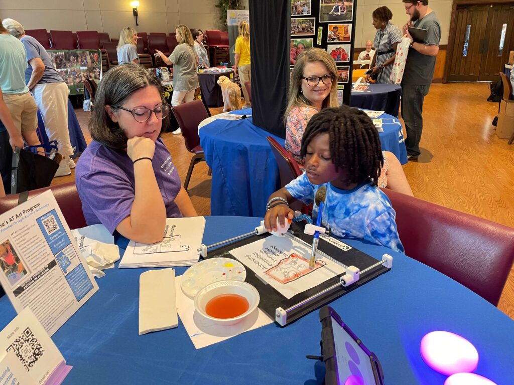 A child works on an art project with Jessica watching at a table covered in art supplies