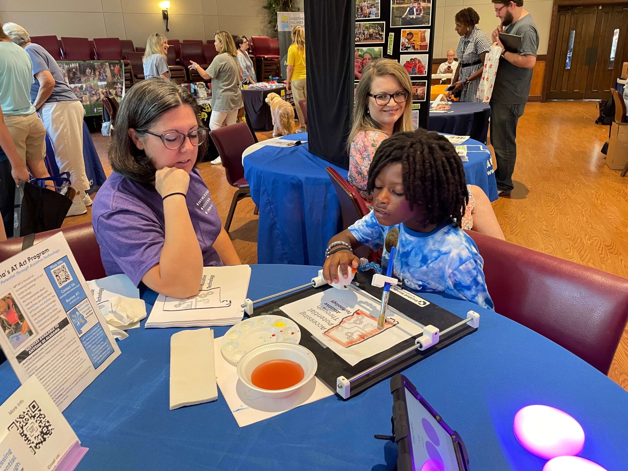 A child works on an art project with Jessica watching at a table covered in art supplies