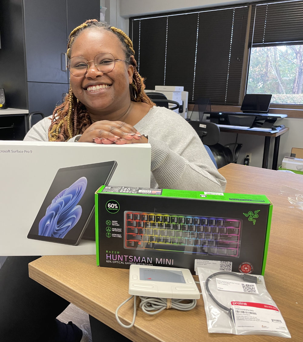 A woman sits at a desk smiling, with a Microsoft Surface Pro 9, a Razer Huntsman Mini keyboard, and other tech items in front of her.