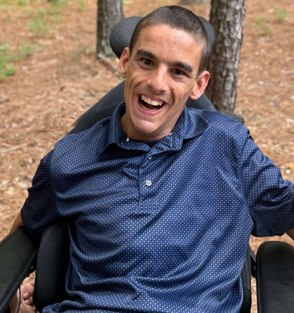 Hank Poore is wearing a blue patterned shirt. He sits in a wheelchair outdoors, smiling at the camera with trees and pine needles in the background.