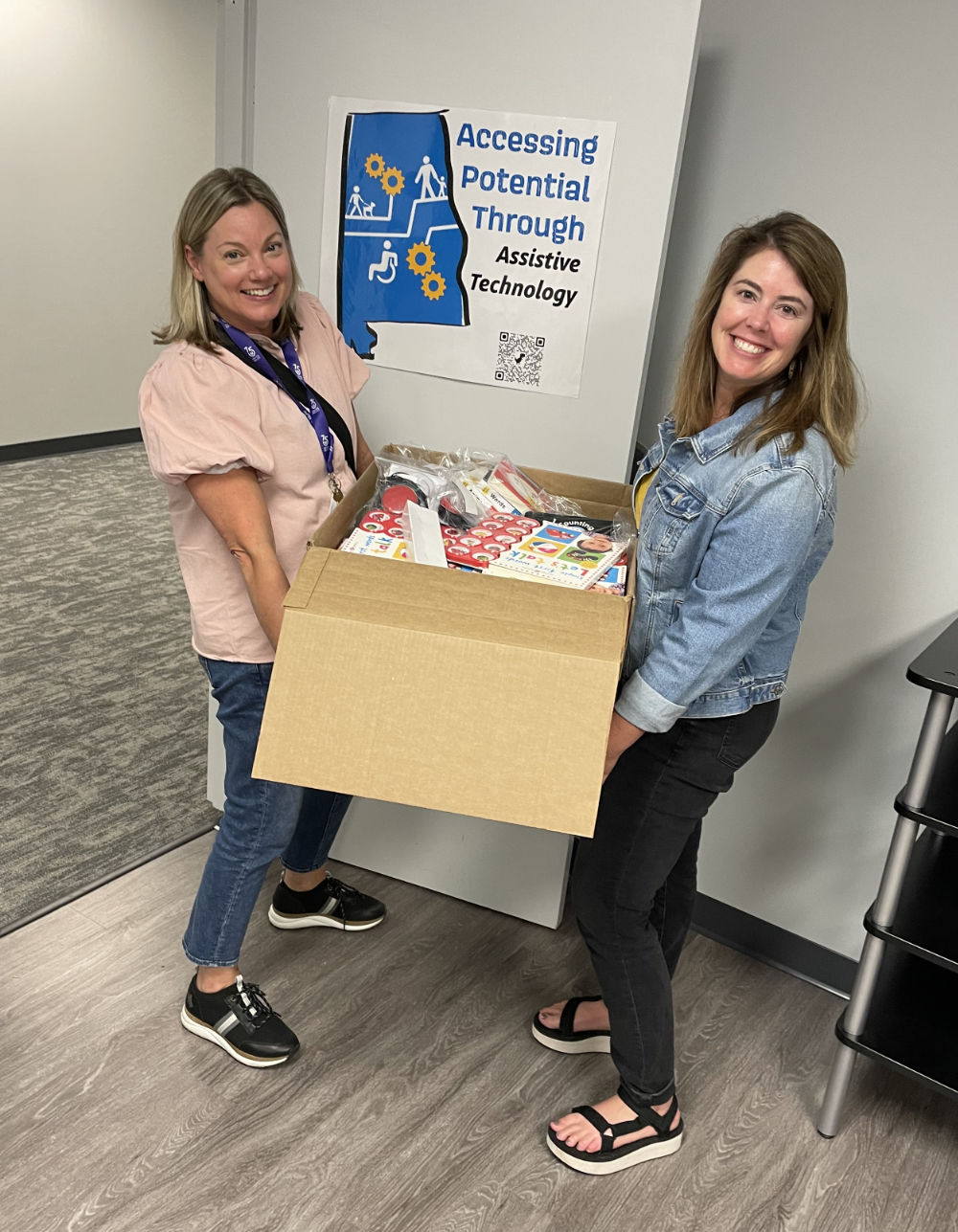 Lynn and Elizabeth from APTAT stand indoors, smiling and holding a large box filled with supplies in front of a sign that reads "Accessing Potential Through Assistive Technology.