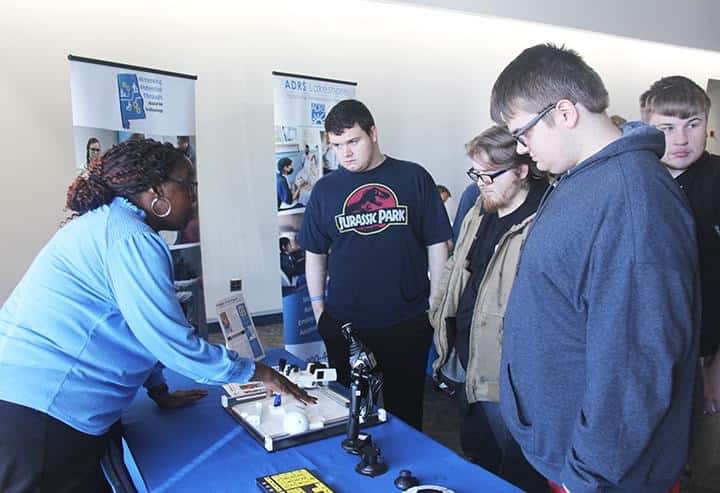 APTAT staff demonstrates an assistive technology at a booth while a group of young men attentively observe.