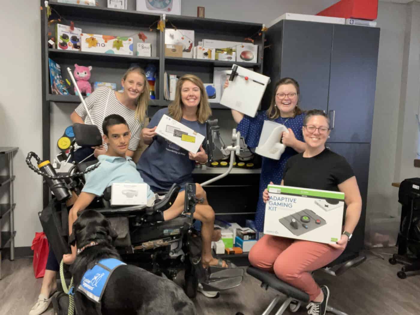 The APTAT team pose indoors holding adaptive gaming kits and devices, smiling at the camera. Shelves with various assistive devices are visible in the background.