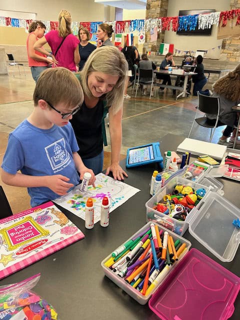 A young boy colors a worksheet with marker stamps at a table covered in art supplies, while a woman stands beside him, smiling.