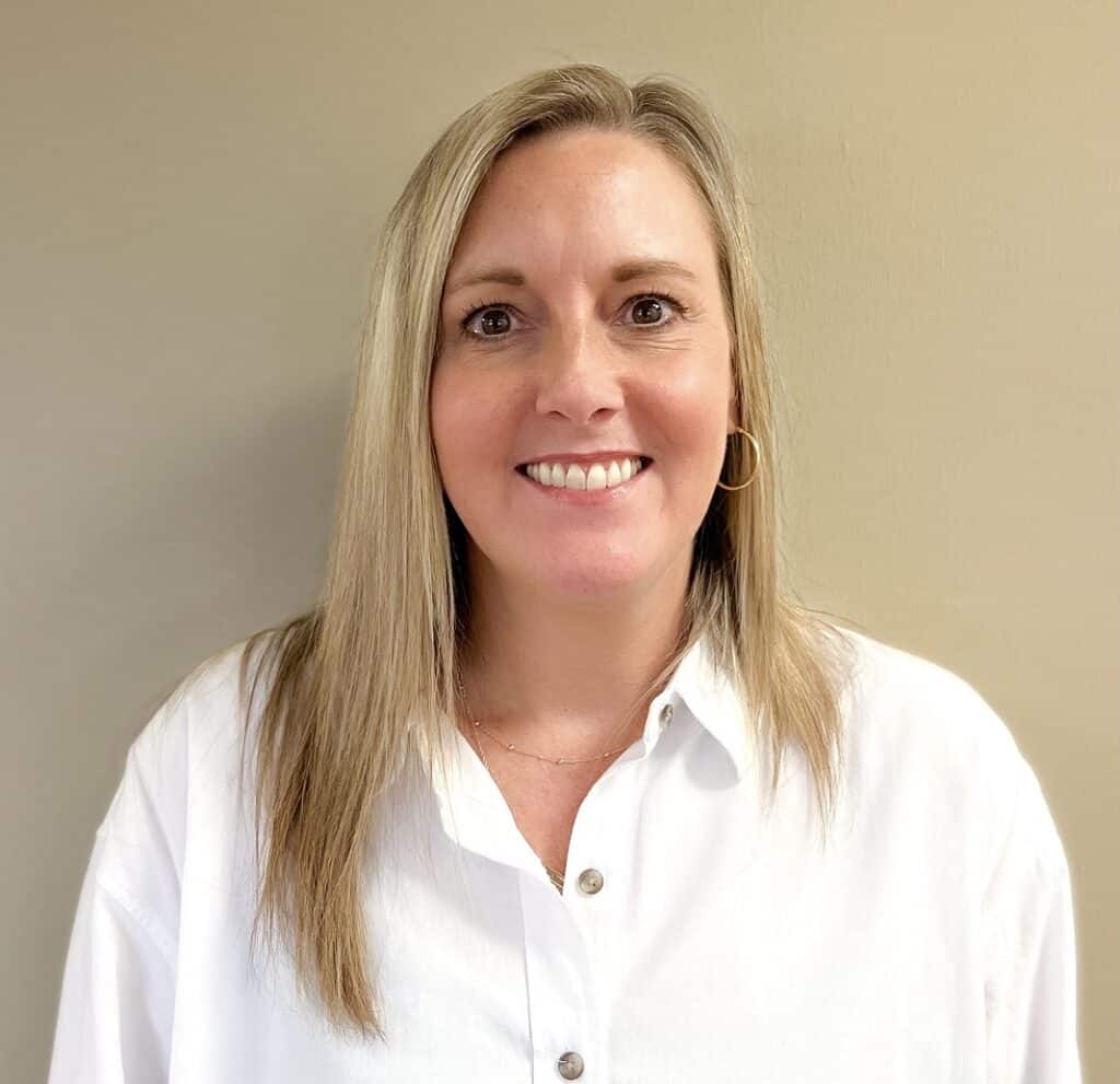 Karen Baggett has straight blonde hair and a white button-up shirt, standing in front of a beige wall, smiling at the camera.