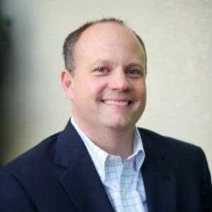 Kevin Braswell is in a dark suit jacket and white checkered shirt smiles at the camera against a neutral background.