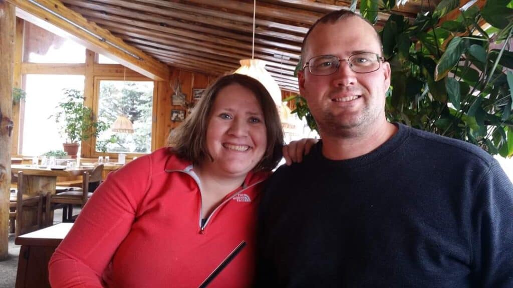 Brinda Dungan and a companion sit together and smile inside a wooden restaurant with large windows and plants.