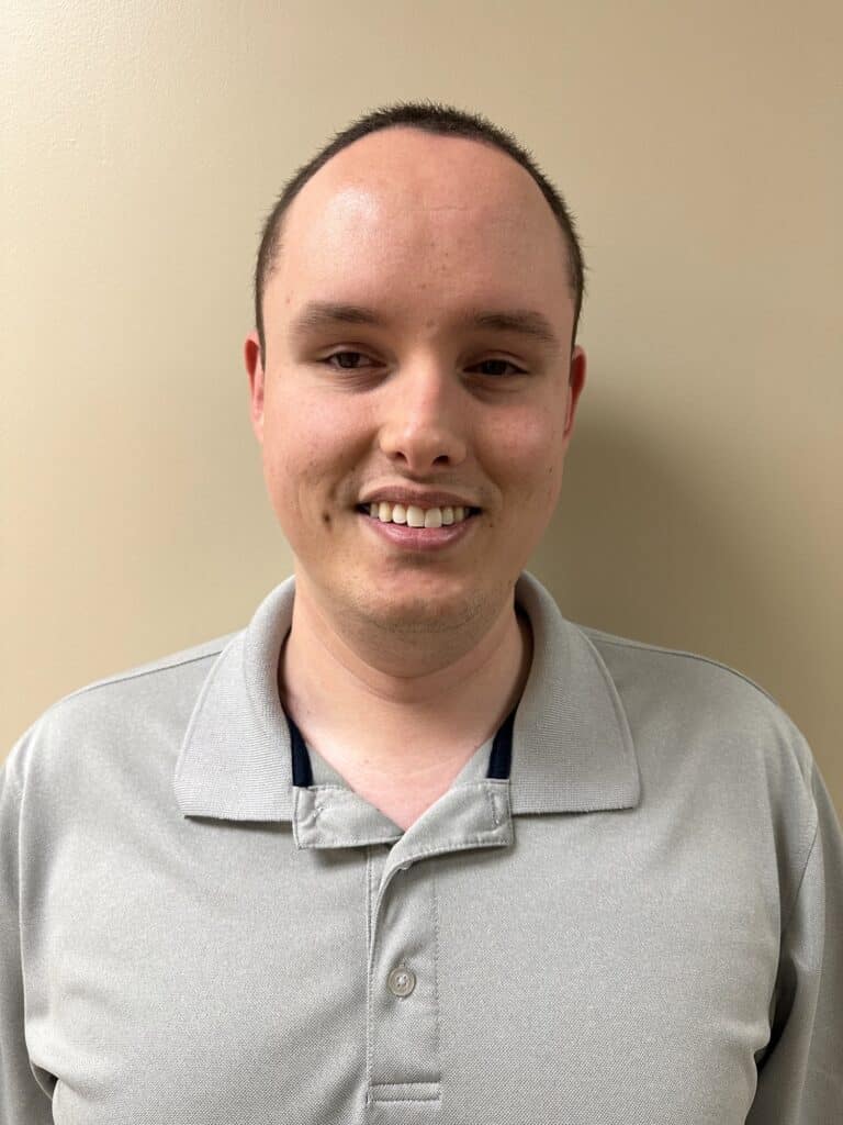 Collin Frase has short hair, wearing a light gray polo shirt, stands in front of a beige wall, smiling at the camera.