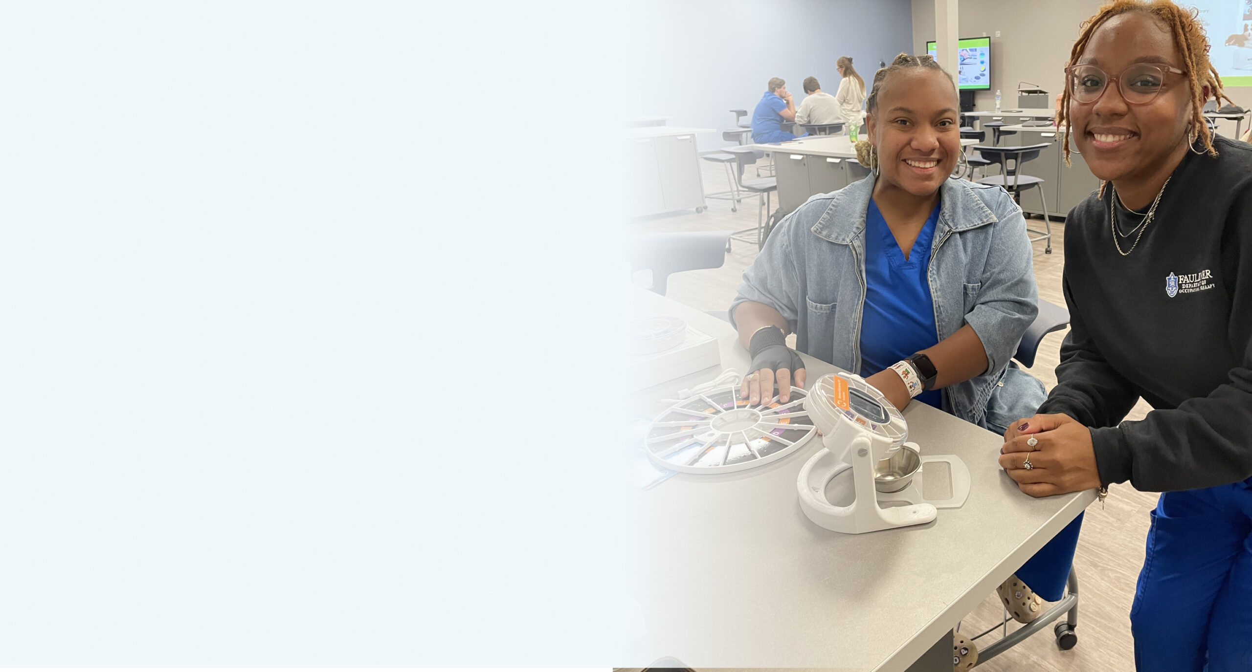 Two women wearing blue uniforms and glasses sit at a table with assistive devices, smiling at the camera.