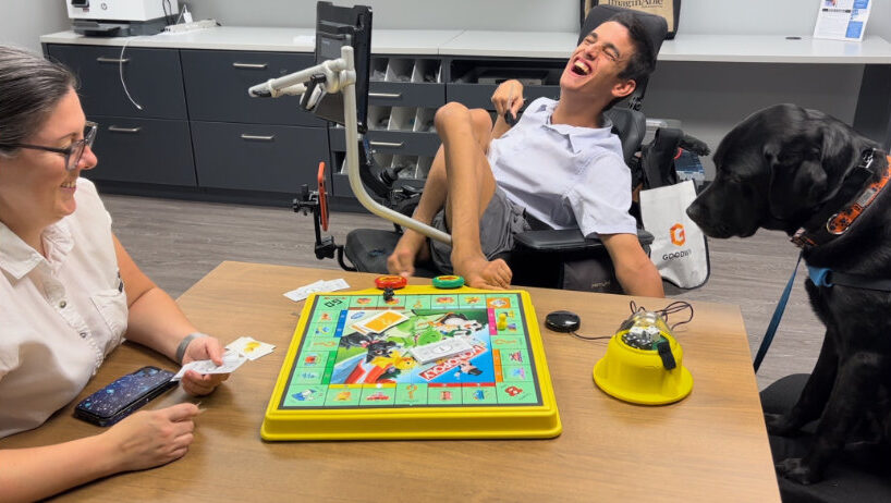 APTAT team members Jessica and Hank with his service dog sit at a table playing a board game. Hank uses a wheelchair and an assistive device to play.