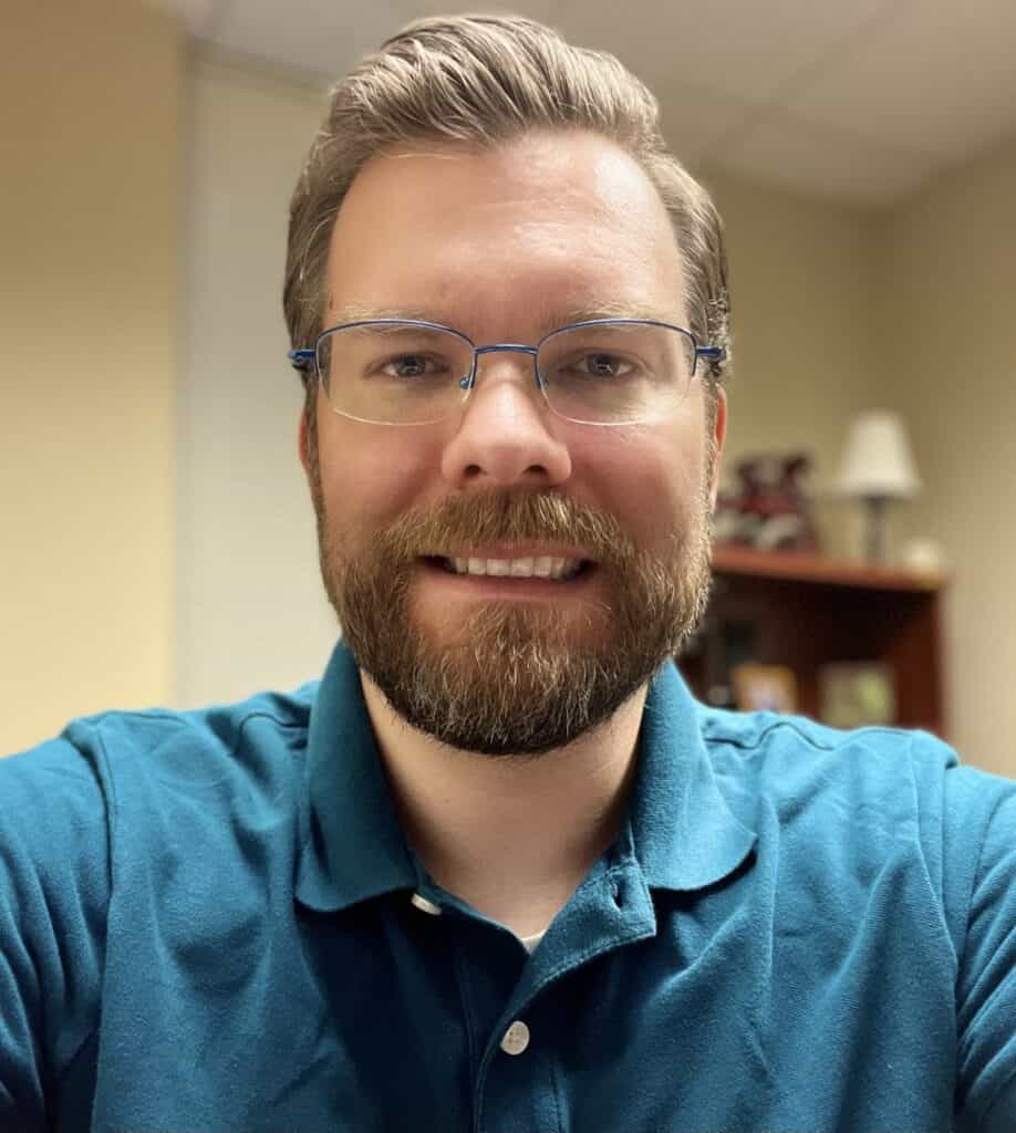 Daniel Lute has a beard, wearing glasses and a teal polo shirt, smiles at the camera in an office setting.