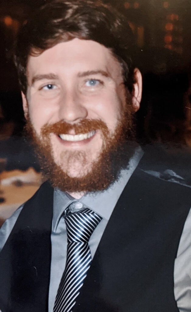 Jason Martin has a brown beard and mustache, wearing a gray shirt, striped tie, and dark vest, smiles at the camera in an indoor setting.
