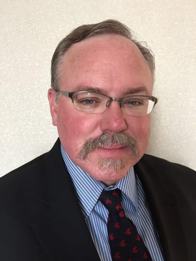Michael Papp has glasses, a mustache, and short hair wearing a dark suit, blue striped shirt, and patterned tie, standing against a light-colored background.