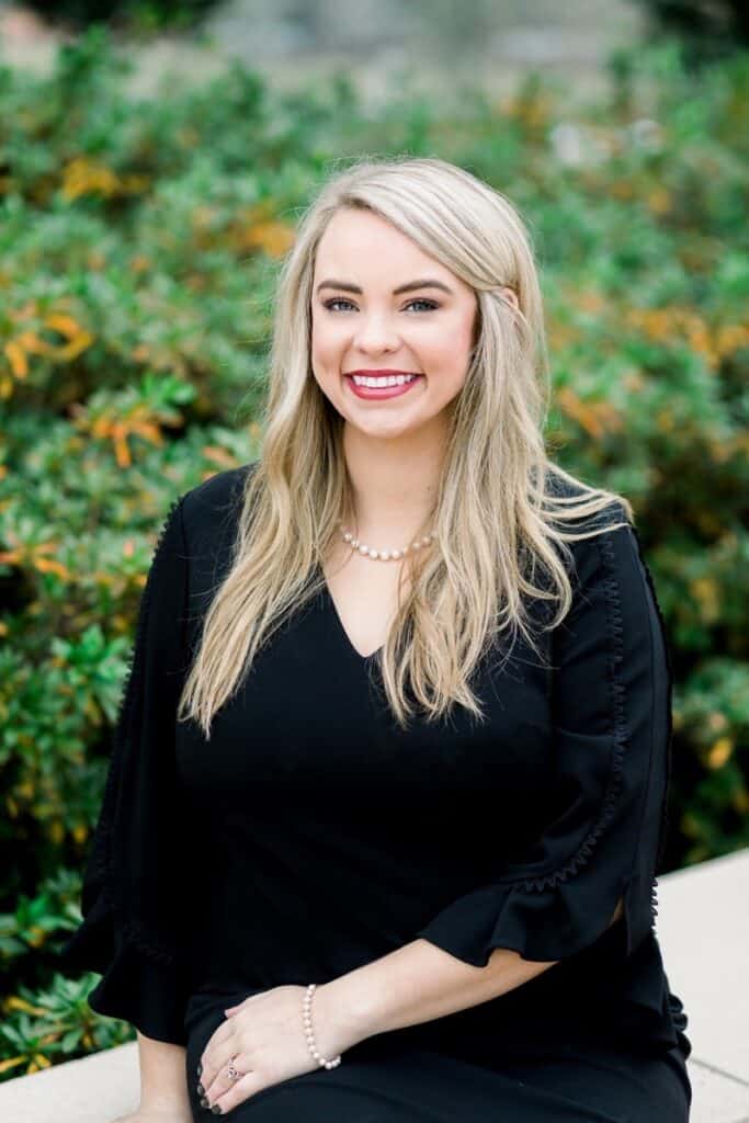 Lauren Perry has long blonde hair, wearing a black dress and pearl jewelry, smiling at the camera.