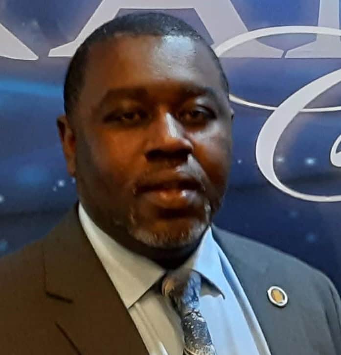 Darryle Powell is wearing a suit and tie, stands in front of a blue background with white text and designs.