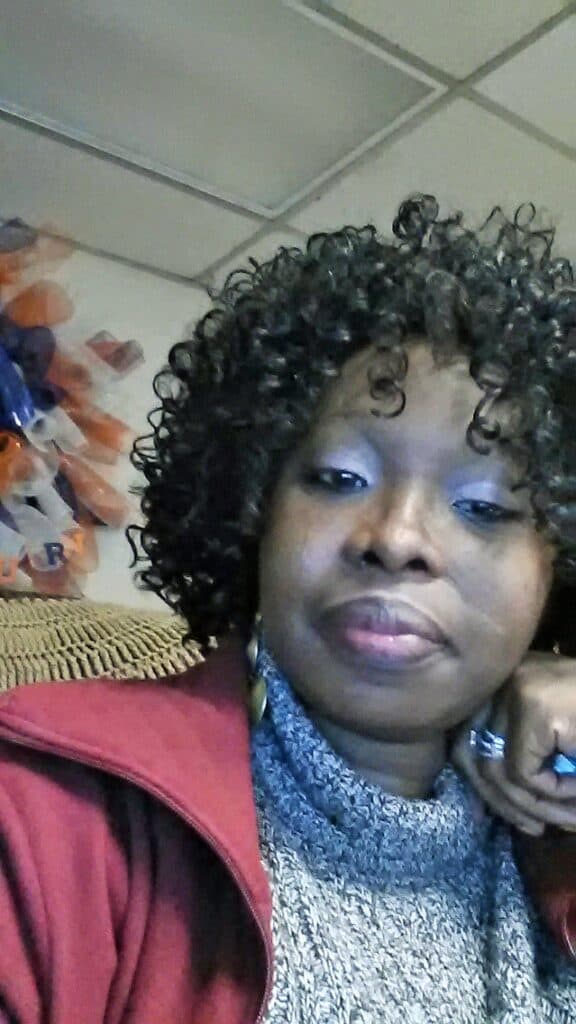 Godilean Powell has curly hair, wearing a gray turtleneck and red jacket, sits indoors, resting her chin on her hand and looking at the camera.