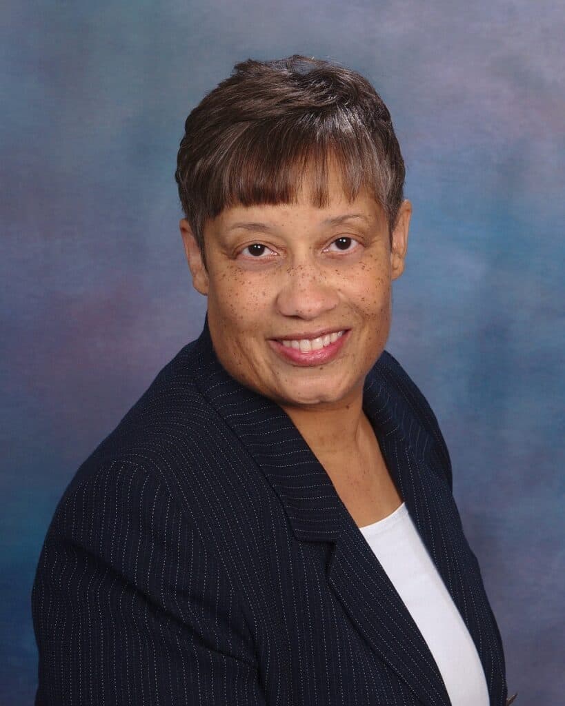 Renee Scruggs has short hair, wearing a dark pinstripe blazer and white shirt, smiling in front of a blue and purple gradient background.